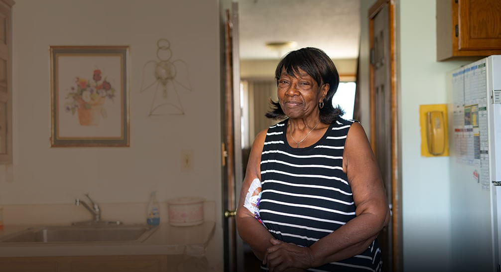 Smiling middle-aged woman standing in her kitchen with a medical patch on her right bicep.