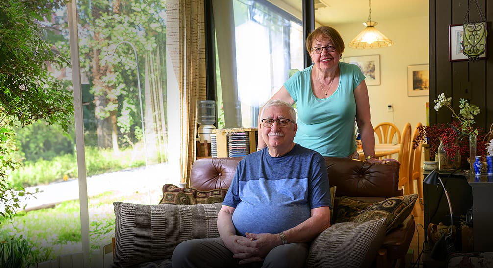 Elderly couple smiling at home, with man sitting on a sofa and woman standing behind.