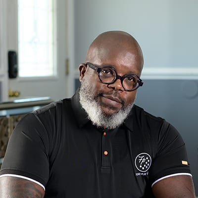 Middle aged African American man sitting on his couch in his living room wearing black circular rimmed glasses.